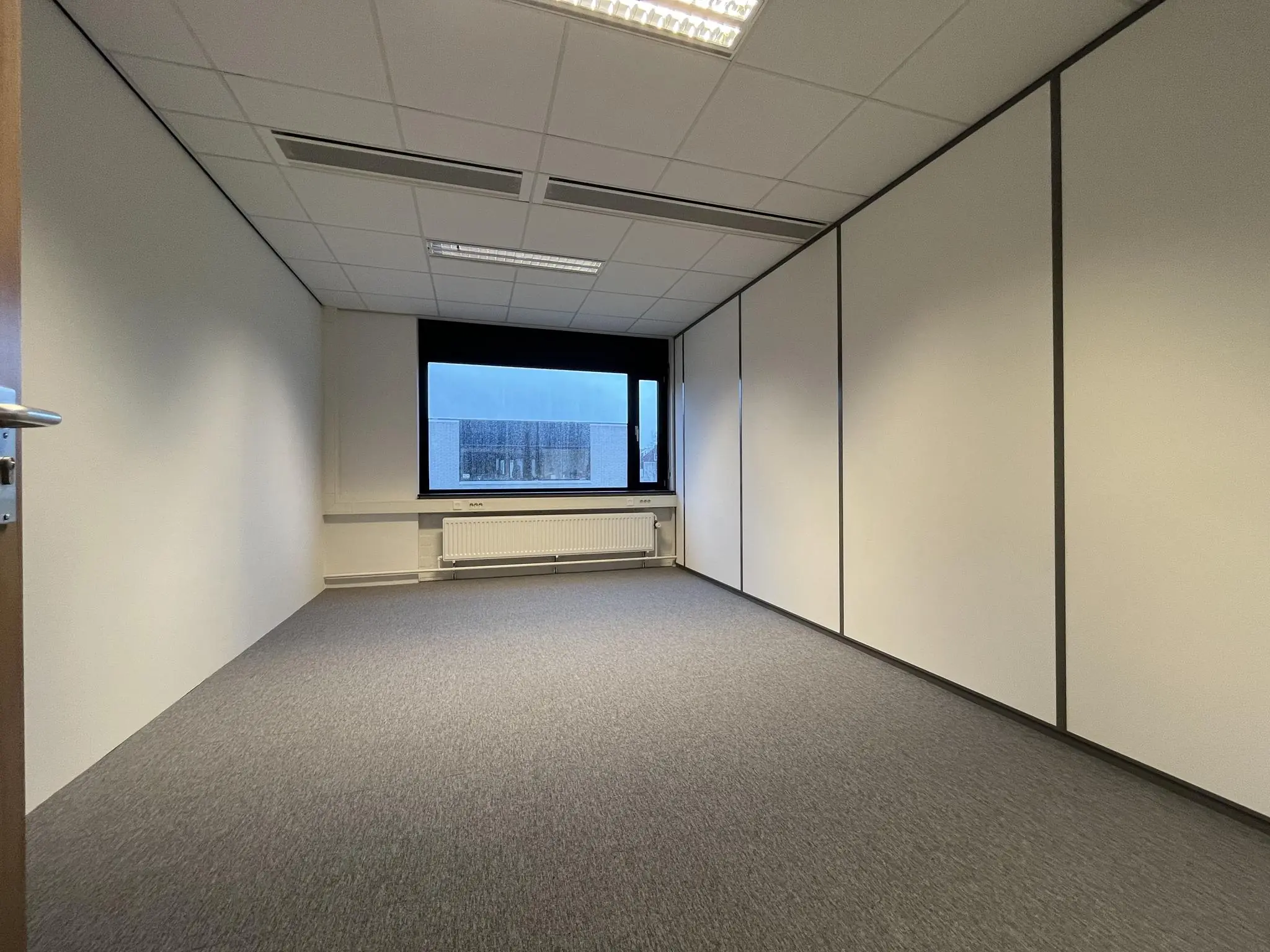 Leeg kantoorruimte met witgesausde wanden, systeemplafond en een raam met uitzicht op een gebouw aan de Burgemeester Verderlaan.